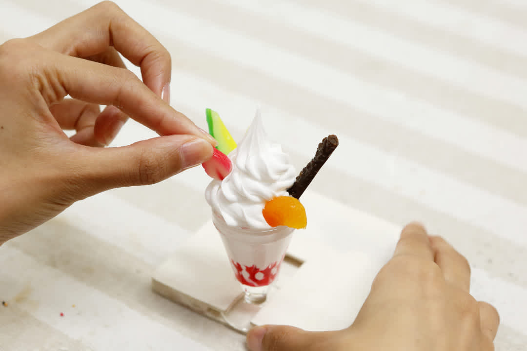 A hand adding a piece of strawberry replica to a small ice-cream sundae model