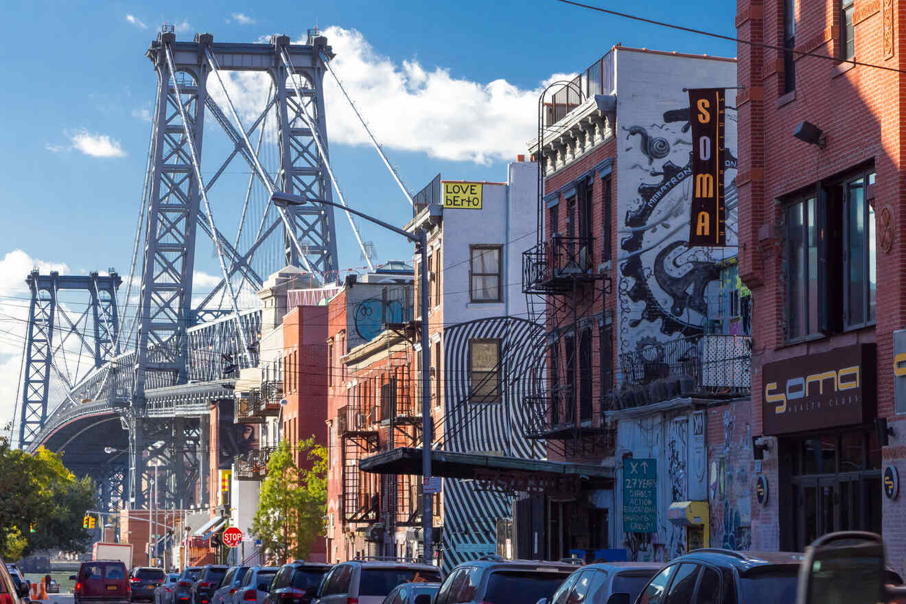 Street scene with buildings and street art beside the Williamsburg Bridge. Traffic moves along the street under a blue sky with scattered clouds.