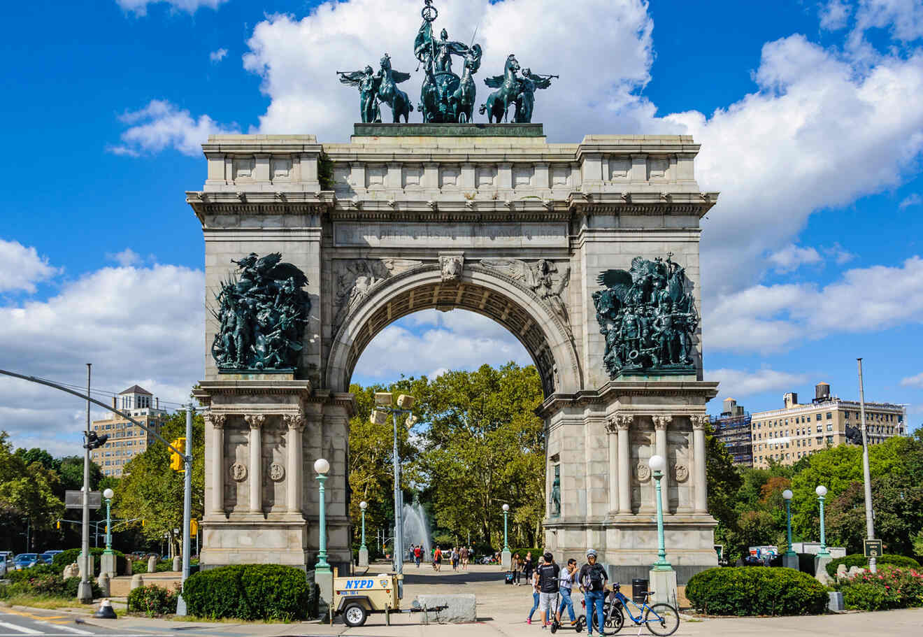 Grand Army Plaza in Brooklyn, featuring the Soldiers' and Sailors' Arch with people walking and biking nearby on a sunny day.
