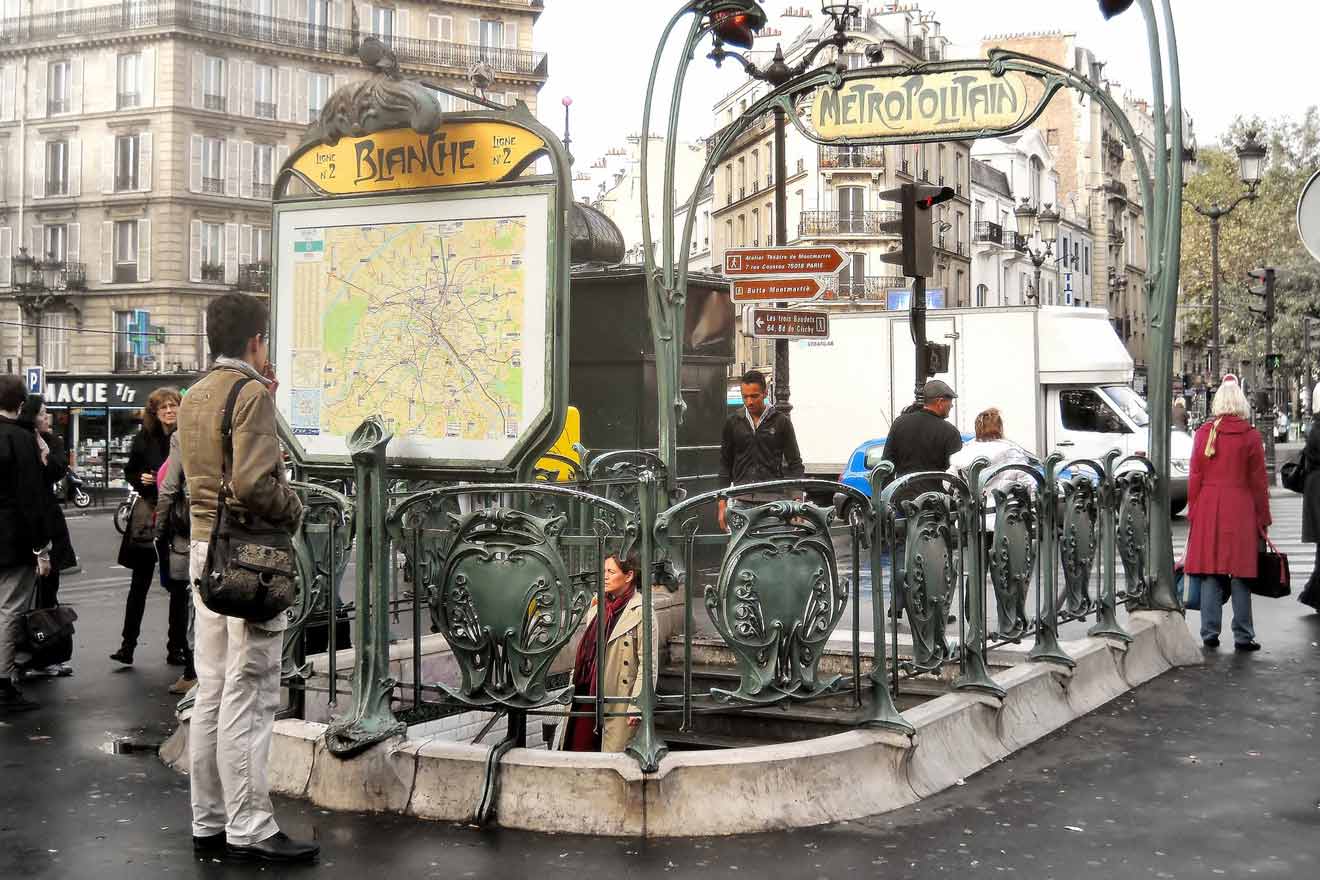 People standing near the Blanche Métro station entrance in Paris, with an Art Nouveau design, and a map display. Buildings and street signs are visible in the background.