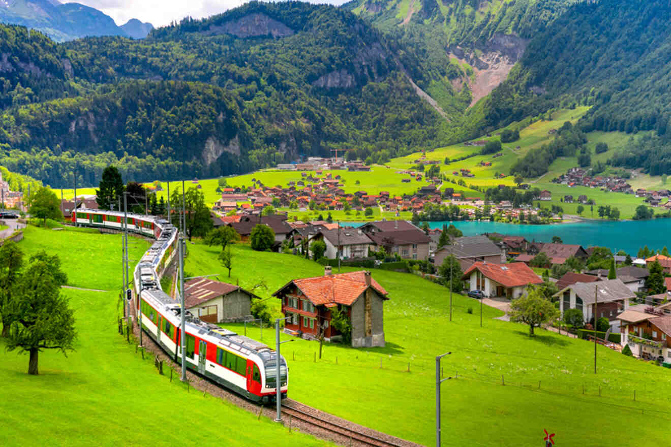 A red and white train passes through a scenic rural landscape in Switzerland with green fields, scattered houses, a small lake, and forested mountains in the background.