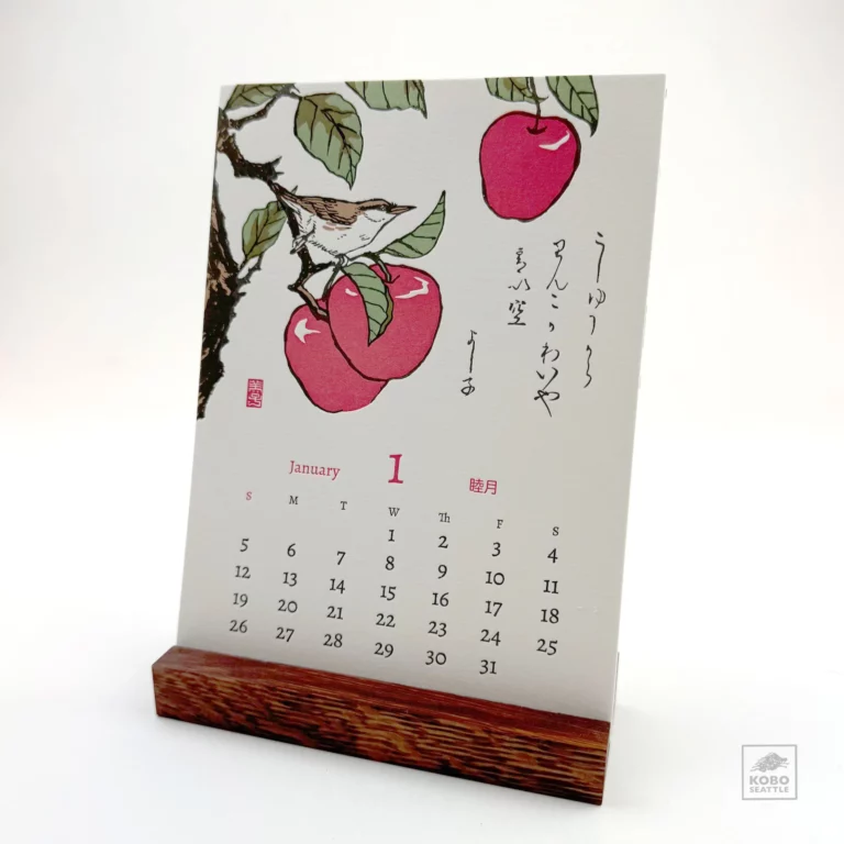 A January calendar page on a wooden stand, featuring a traditional Japanese artwork of a bird perched on a branch with red fruit. The artwork includes Japanese characters, and the month's dates are displayed below.